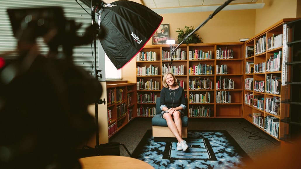 Lachende vrouw zittend in een kamer vol boeken met een camera op zich gericht voor het maken van een documentaire.