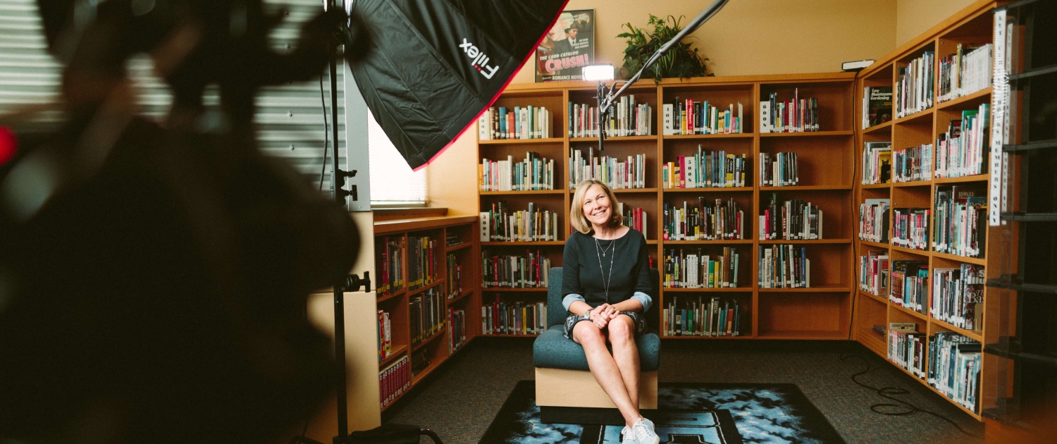 Lachende vrouw zittend in een kamer vol boeken met een camera op zich gericht voor het maken van een documentaire.